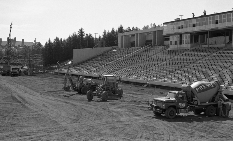 Black and white photograph of heavy machinery and construction equipment on the field of a concrete, open air stadium.