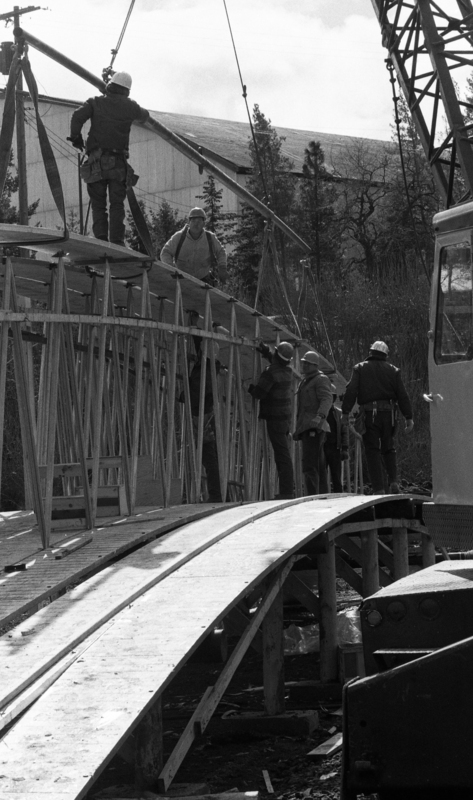 Black and white photograph of several men standing on a wooden arch that sits on the ground in a construction site.