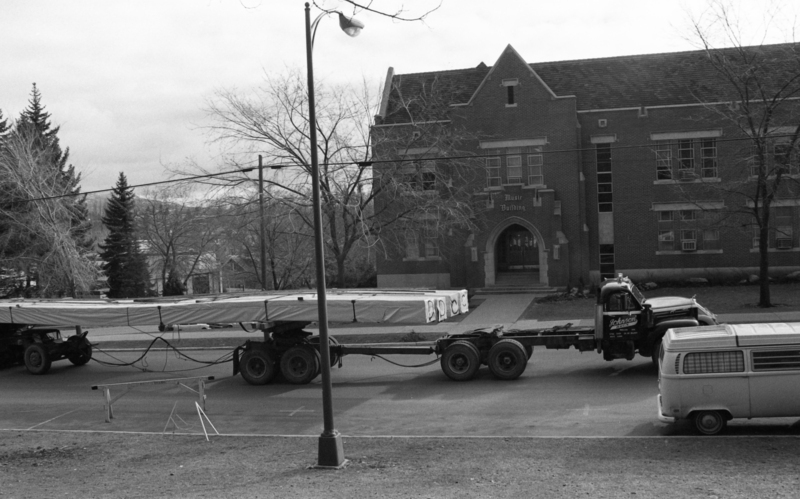 Black and white photograph of a truck towing a trailer loaded with long, thin cargo, driving through city streets. In the foreground is a Volkswagen bus, and in the background is a building labeled "Music Building".