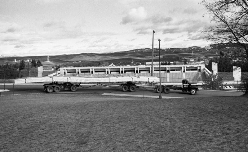 Black and white photograph of a truck driving by an open air stadium, loaded with long, thin cargo.