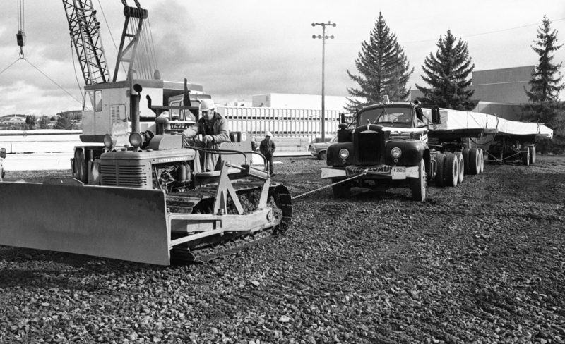 Black and white photograph of a man driving a tractor that is chained to a truck that is loaded with long, thin cargo, through a construction site.