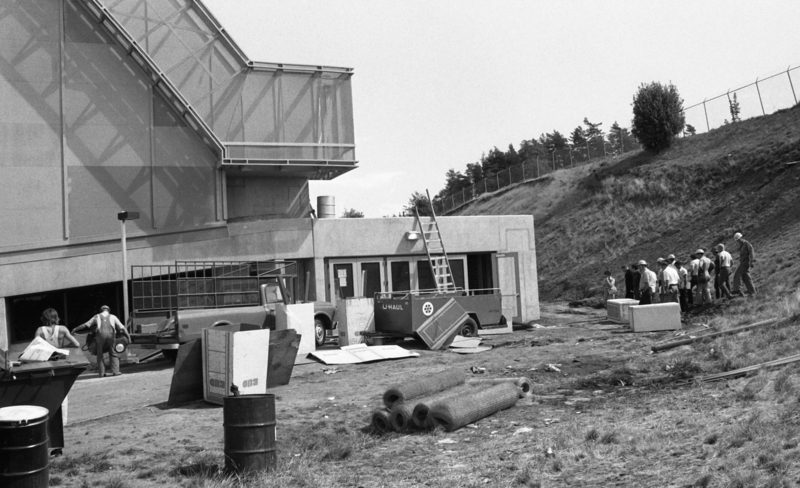 Black and white photograph of a group of people standing on a hillside next to a concrete building.