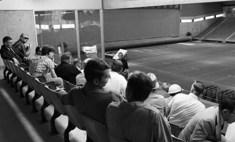 Black and white photograph of a group of people sitting in the luxury box of an enclosed stadium.