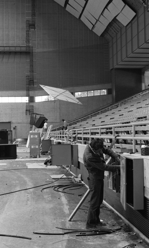 Black and white photograph of a man working on a a metal box attached to an end wall in an enclosed stadium. In the background, a ceiling tile is being raised into place.