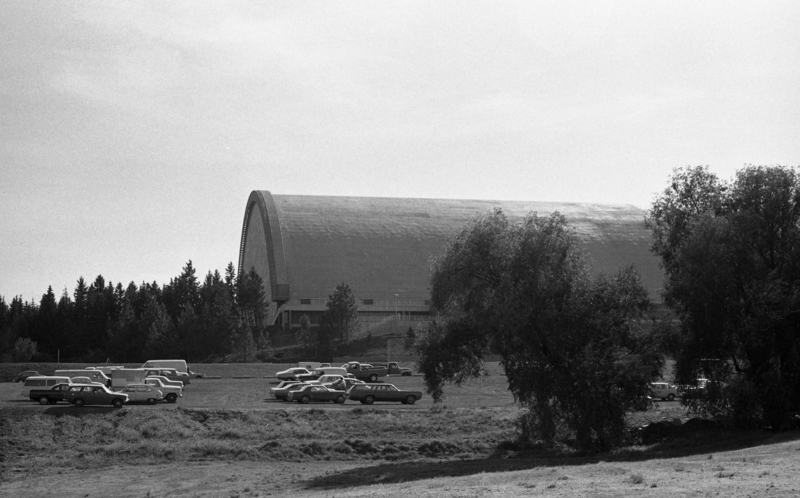 Black and white photograph of a building with an arched roof. In the foreground are cars in a parking lot.