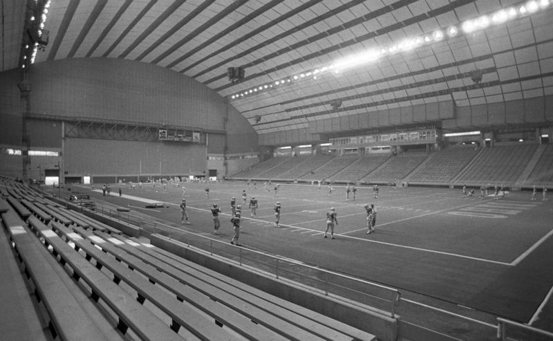 Black and white photographs of the interior of an enclosed stadium. Football players stand around the football field. The stands are empty.