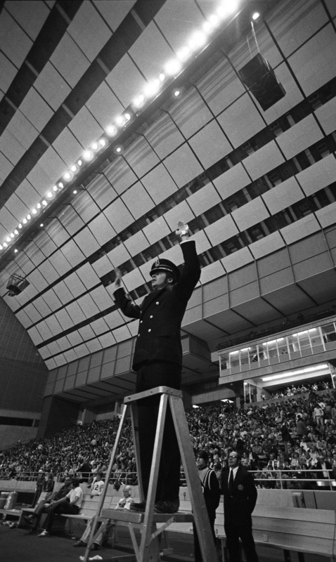 Black and white photograph of a man standing on a short ladder, his arms raised. In the background, people sit in stands.