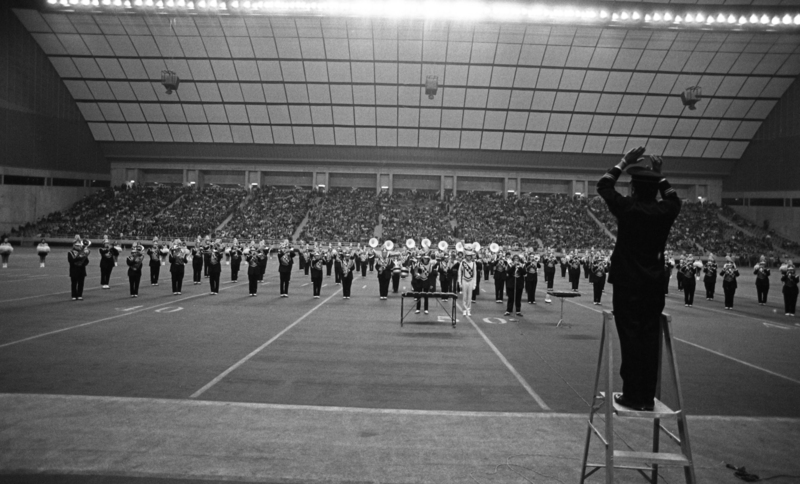 Black and white photograph of a marching band on a football field, being conducted by a person standing on a stepstool in the foreground. In the background are stands crowded with people.