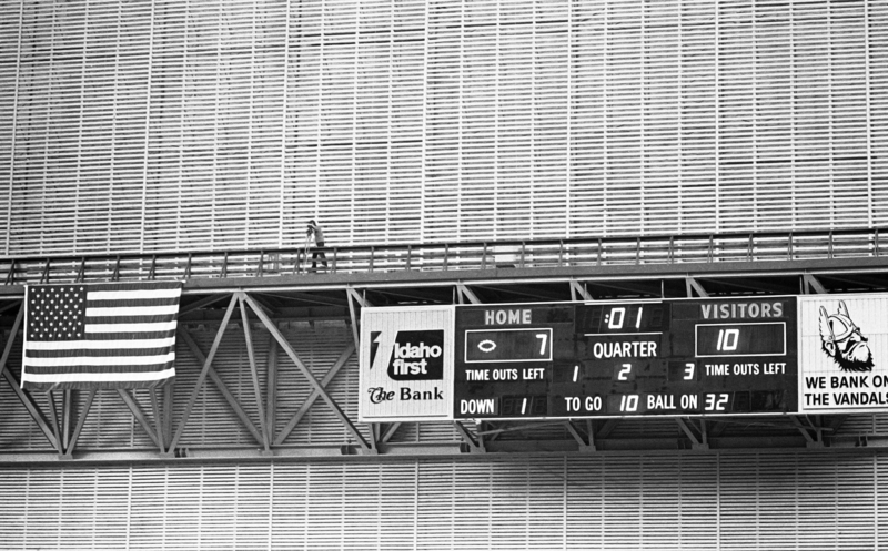 Black and white photograph of a photographer standing on the catwalk of a building. Below the catwalk is a scoreboard and a US flag.
