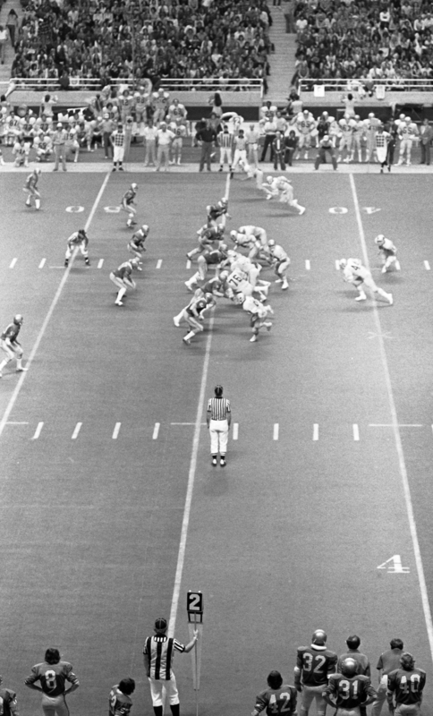 Black and white photograph of football players running at each other on a football field. In the foreground are referees and more football players. In the background are spectators in stands.