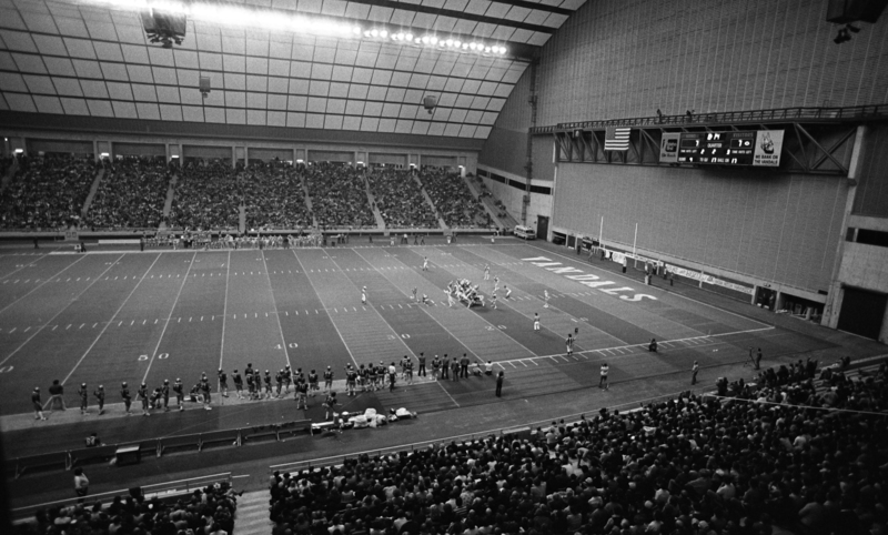 Black and white photograph of a football game taking place in an enclosed stadium.