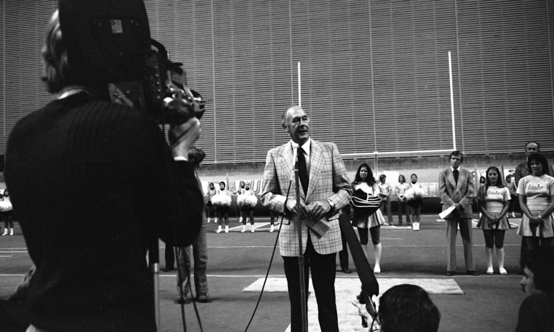 Black and white photograph of a man speaking into a microphone. In the foreground is a person holding a film camera. In the background are more people and football goalpost.