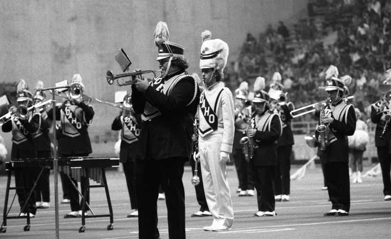 Black and white photograph of people dressed in marching band uniforms and playing wind instruments. In the background are stadium stands crowded with people.