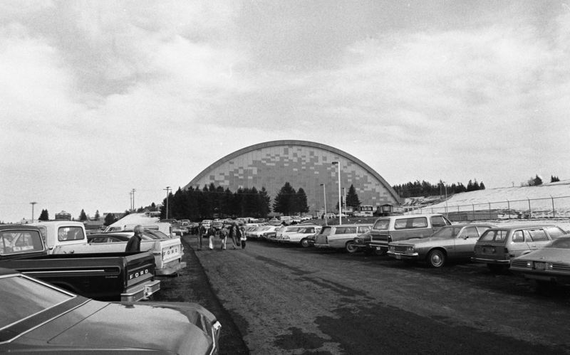 Black and white photograph of cars in a parking lot. In the background is a building with an arched roof.