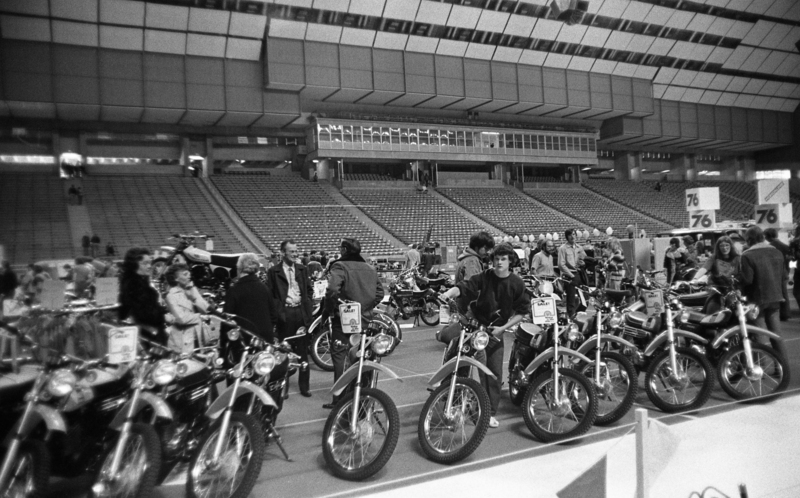 Black and white photograph of people walking by motorbikes parked on the field of an enclosed stadium.