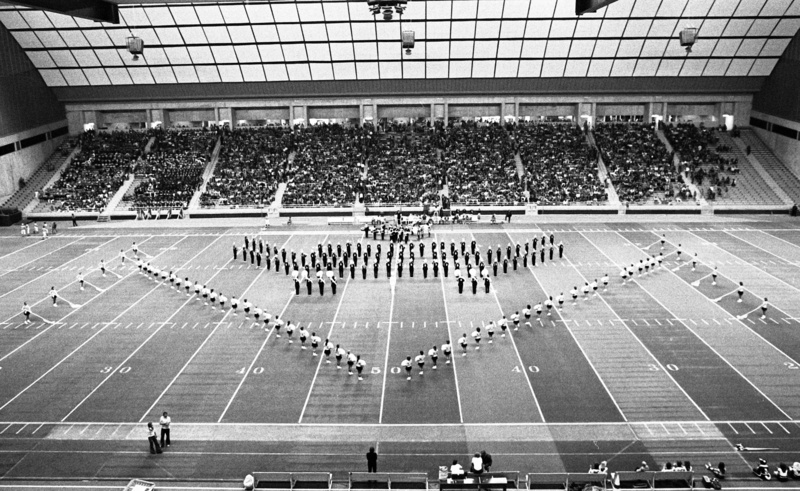 Black and white photograph of a marching band in formation on a football field. In the background are stands filled with people.