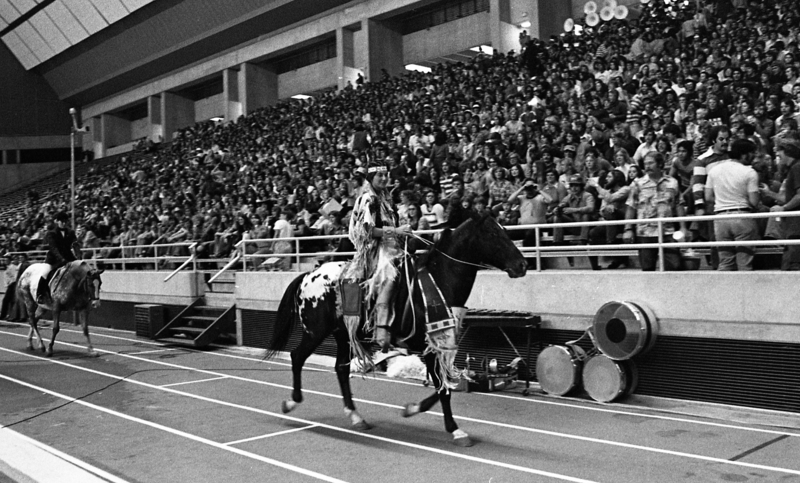 Black and white photograph of two women riding horses along an indoor running track. In the background are people sitting in stadium stands.