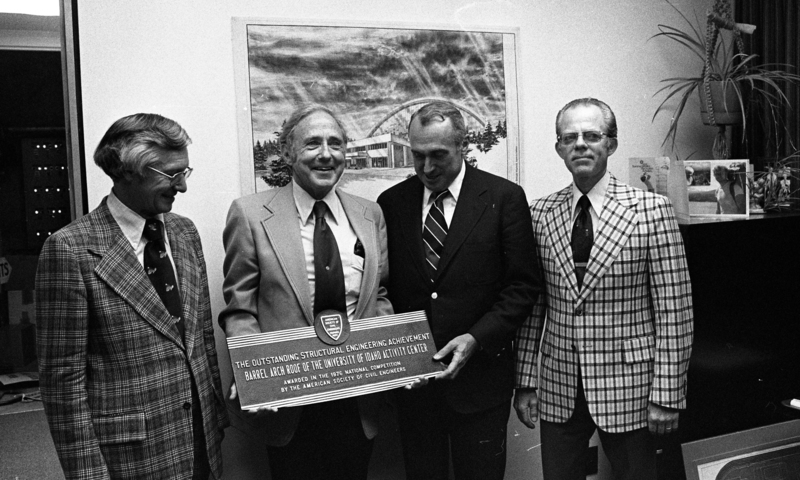 Black and white photograph of four men standing in an office. One of them holds a plaque that reads "The Outstanding Structural Engineering Achievement Barrel Arch Roof of the University of Idaho Activity Center Awarded in the 1976 National Competition by the American Society of Civil Engineers"