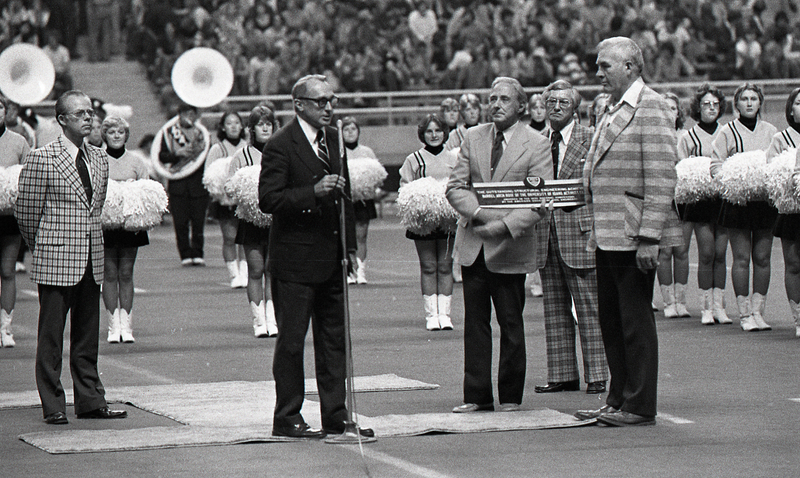 Black and white photograph of several people standing on a football field, one of whom is speaking into a microphone. Another holds a plaque.