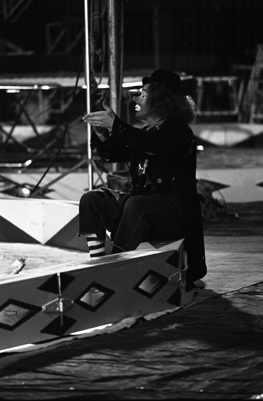 Black and white photograph of a person dressed as a clown sitting on the side of a circus ring.