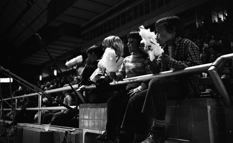 Black and white photograph of four children sitting in the front row of stadium stands and eating cotton candy.