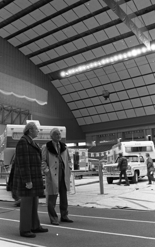 Black and white photograph of two men standing on an indoor track in an enclosed stadium. In the background is a truck, campers, and people milling around.