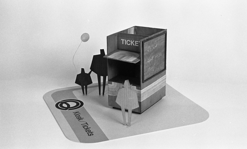 Black and white photograph of a miniature mockup of a ticket booth surrounded by abstract human-shaped cutouts.