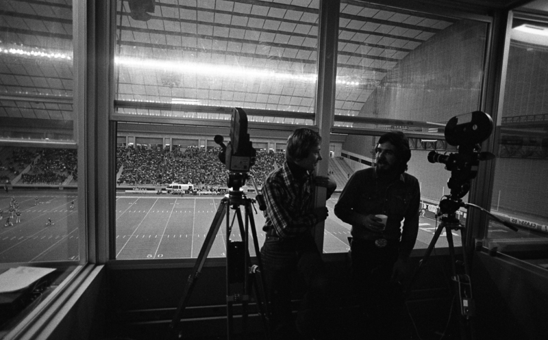 Black and white photograph of two people standing beside film cameras. In the background is an enclosed football stadium.