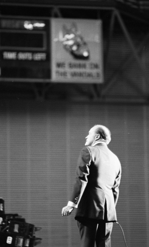 Black and white photograph of a man in a suit, bathed in light. In the background is a scoreboard.
