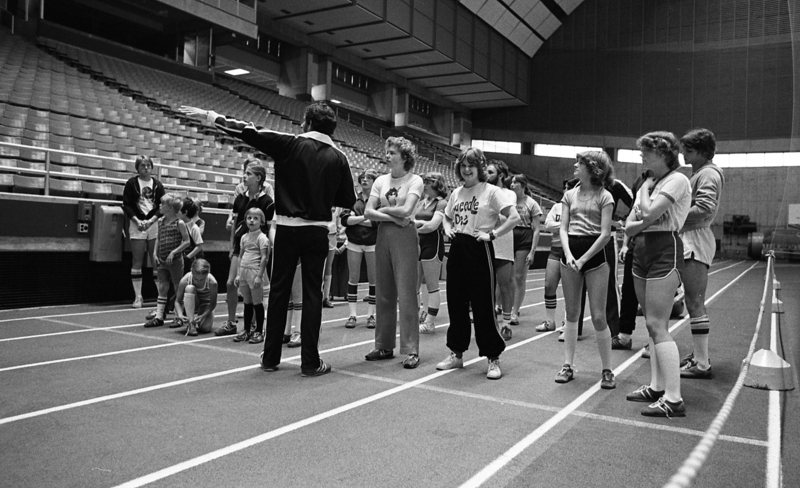 Black and white photograph of a group of people in running gear on a running track in an enclosed stadium.