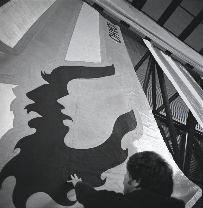 Black and white photograph of several banners with the names of universities hanging along the inside wall of a stadium.