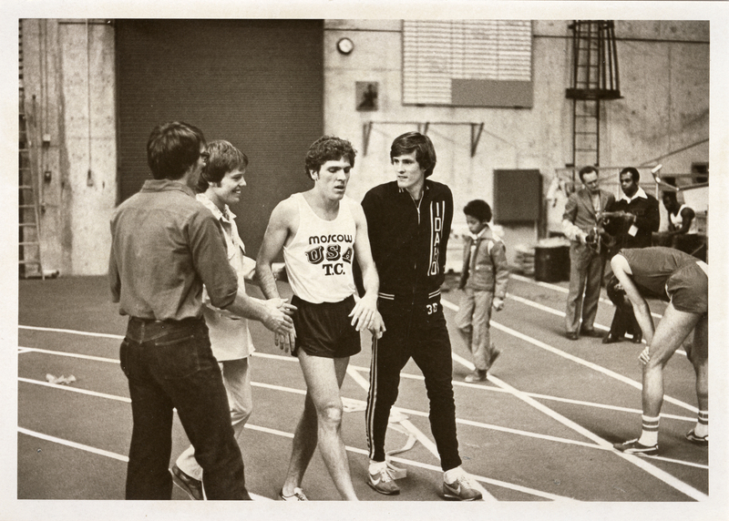 Black and white photograph of an athlete walking along an indoor track and interacting with other people.
