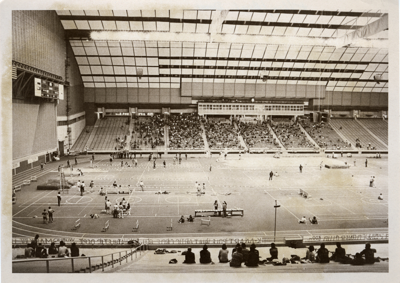 Black and white photograph of an enclosed stadium. Various activity setups are scattered around the field, and people sit in the stands in the background and foreground.