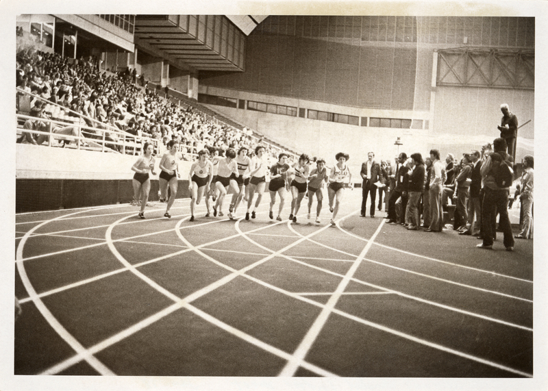 Black and white photograph of a group of women running on an indoor track in an enclosed stadium. People sit in the stands on the left.