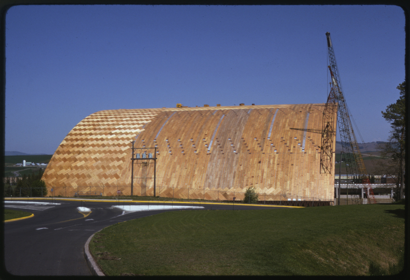 Color photograph of a building under construction, with a crane at one end. The building has an arched wooden roof.