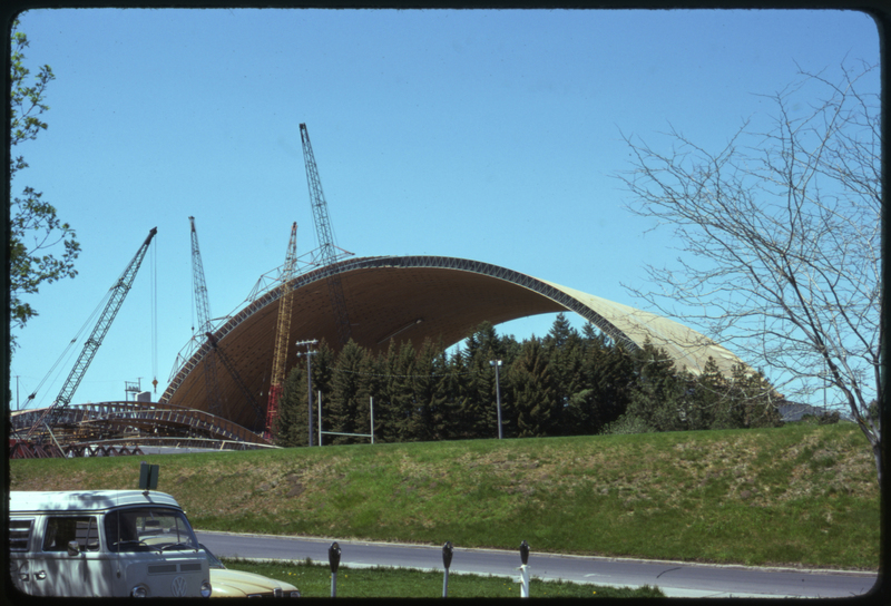 Color photograph of an arched building under construction, surrounded by cranes. A Volkswagen bus appears in the foreground.