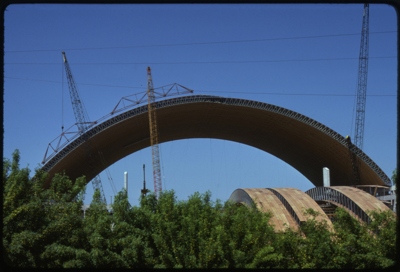 Color photograph of an arched wooden roof under construction, surrounded by construction cranes. In the foreground are more wooden arches and some trees.