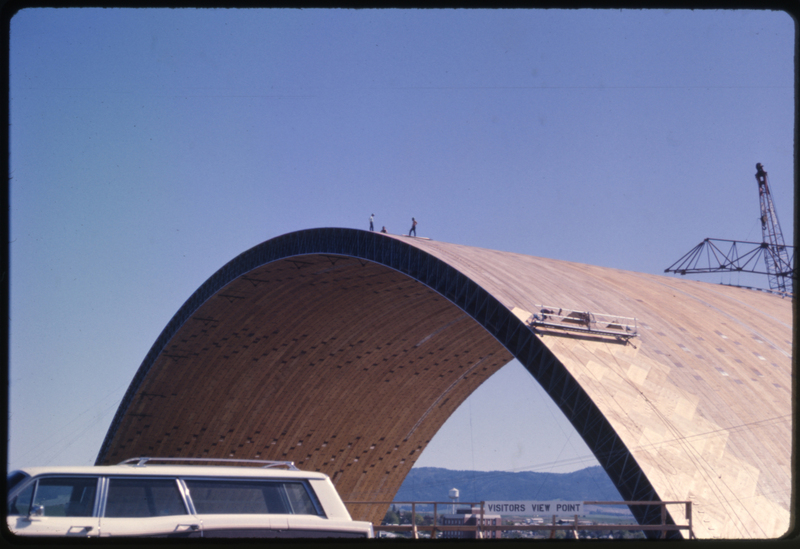 Color photograph of an arched wooden roof under construction. In the foreground is a station wagon.