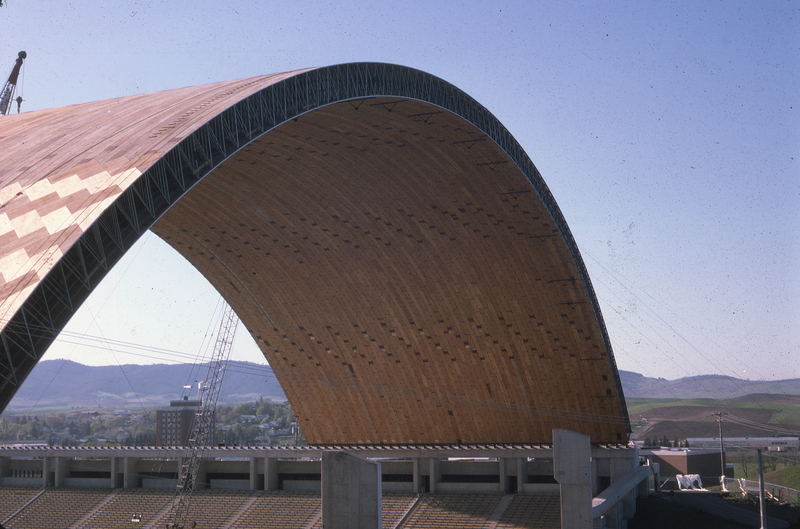 Color photograph of a building with an arched wooden roof under construction. In the background are gently rolling hills.
