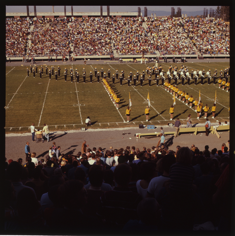 Color photograph of a marching band and cheerleaders in formation on a football field. stands crowded with spectators are in the foreground and background.