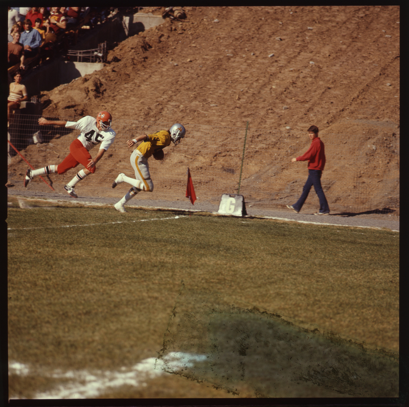 Color photograph of two football players and a bystander on a football field next to a dirt hillside.