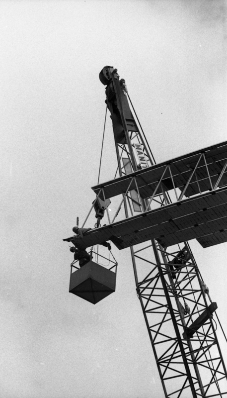 Black and white photograph of two men working on a wooden roof arch, one in a bucket suspended from a crane and the other lying on the roof arch.