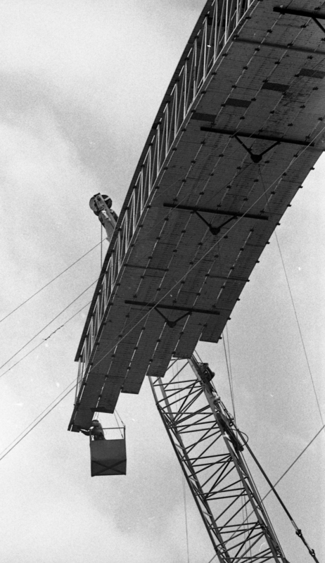 Black and white photograph of one end of a wooden arch rising into the air. A worker in a bucket suspended by a crane works at the end of the arch.