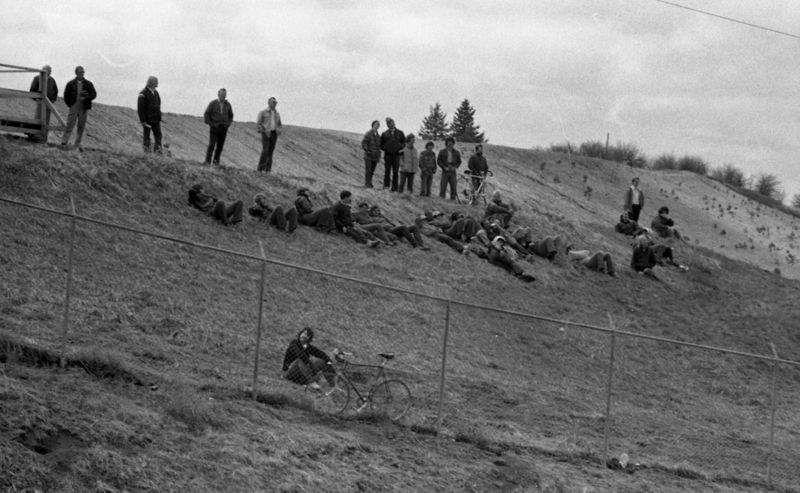 Black and white photograph of people standing or lying on a grassy hillside behind a chain link fence.