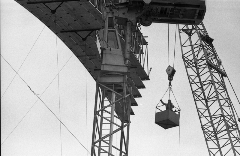 Black and white photograph of a person standing in a bucket suspended from a crane, approaching a wooden arch of an arched roof under construction.