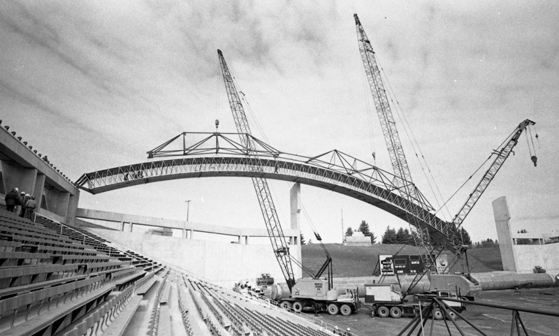 Black and white photograph of a wooden arch being maneuvered by construction cranes in an open air stadium.