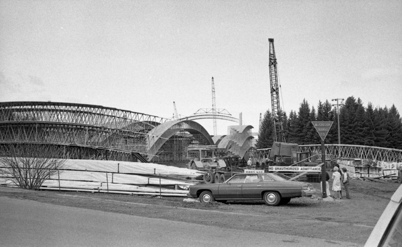 Black and white photograph of a car parked next to a construction site filled with wooden arches mounted on steel trusses.
