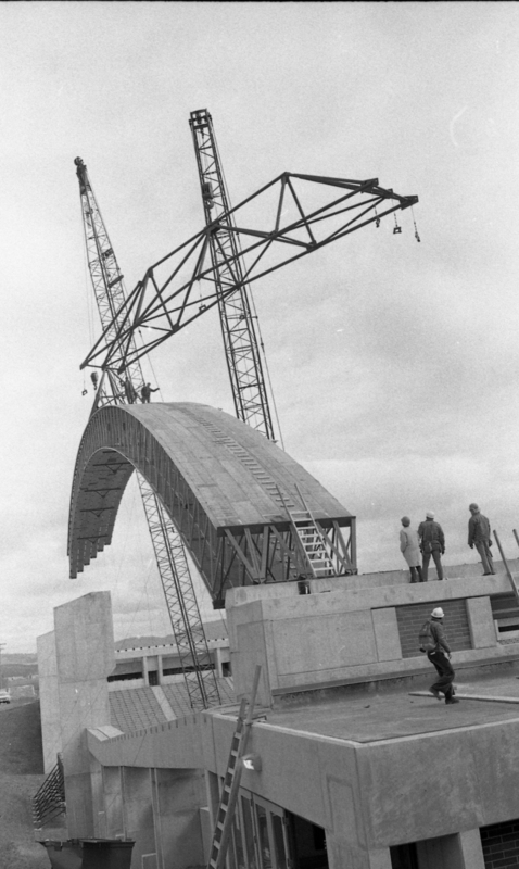 Black and white photograph of a wooden arch being maneuvered by a construction crane. Several people stand on the concrete wall to which the arch will be attached.