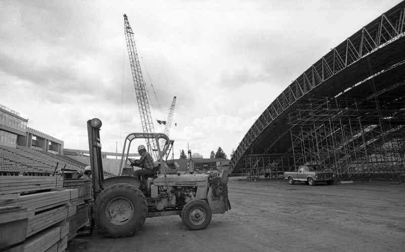 Black and white photograph of a man operating a forklift to transport wooden boards. In the background are long wooden arches and a truck.