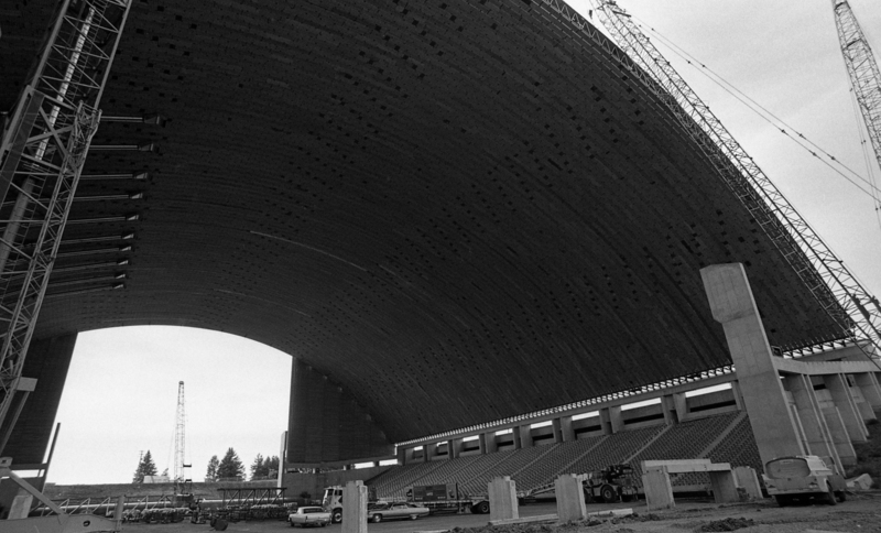 Black and white photograph of an arched roof under construction. Cars and heavy machinery sit on the ground below.